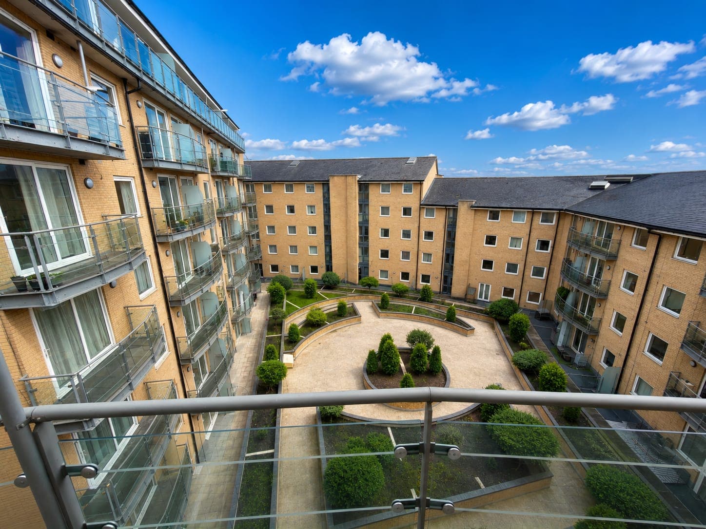 Modern apartment complex featuring spacious communal courtyard with manicured gardens, contemporary brick architecture, and peaceful outdoor seating areas surrounded by well-maintained landscaping.
